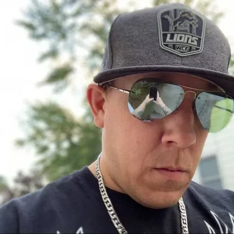 Man in Lions Cap and Aviator Sunglasses with Serious Expression Man wearing a dark Lions cap, aviator sunglasses, and a silver chain, with a serious expression while standing outdoors