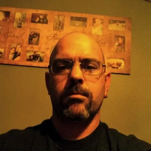 Man with Glasses and Goatee in Front of Family Photo Collage Bald man with glasses and a goatee looking serious in a dimly lit room with a family photo collage on the wall behind him