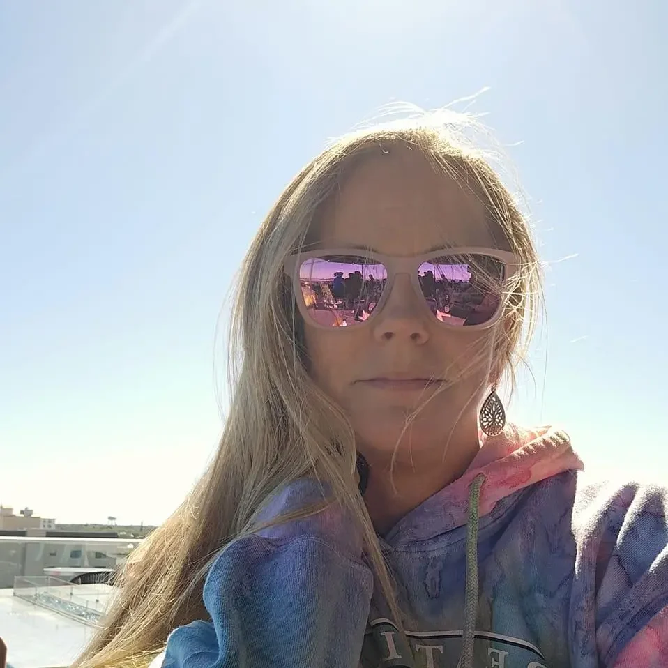 Blonde Woman in Pink Sunglasses and Tie-Dye Hoodie on a Sunny Day Blonde woman wearing pink reflective sunglasses and a tie-dye hoodie, standing outdoors on a sunny day with a bright blue sky in the background.